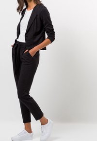 Blazer noir sur mesure à un seul bouton, associé à un pantalon taille élastique avec une ceinture en tissu assorti, complété par des baskets blanches.