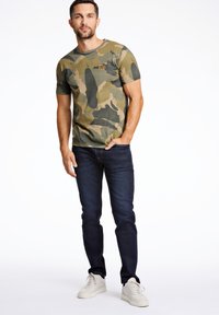 Camiseta de manga corta con estampado de camuflaje en verde y beige, combinada con jeans azul oscuro y zapatillas blancas. Presenta un cuello redondo y un corte ajustado.