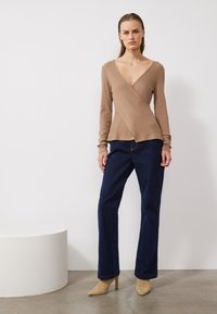 Beige gerippter Wickel-Oberteil mit langen Ärmeln, tiefem V-Ausschnitt, kombiniert mit dunkelblauen geraden Jeans und hellen High Heels.