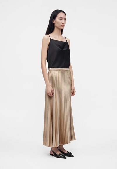 Femme debout portant un haut noir sans manches, une jupe midi plissée beige et des ballerines noires pointues avec détail de boucle sur un fond uni.