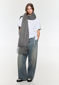 Grauer Strickschal mit Fransen, über einem weißen T-Shirt getragen. Das Model trägt lockere blaue Jeans und schwarze Schuhe und posiert vor einem neutralen Hintergrund.