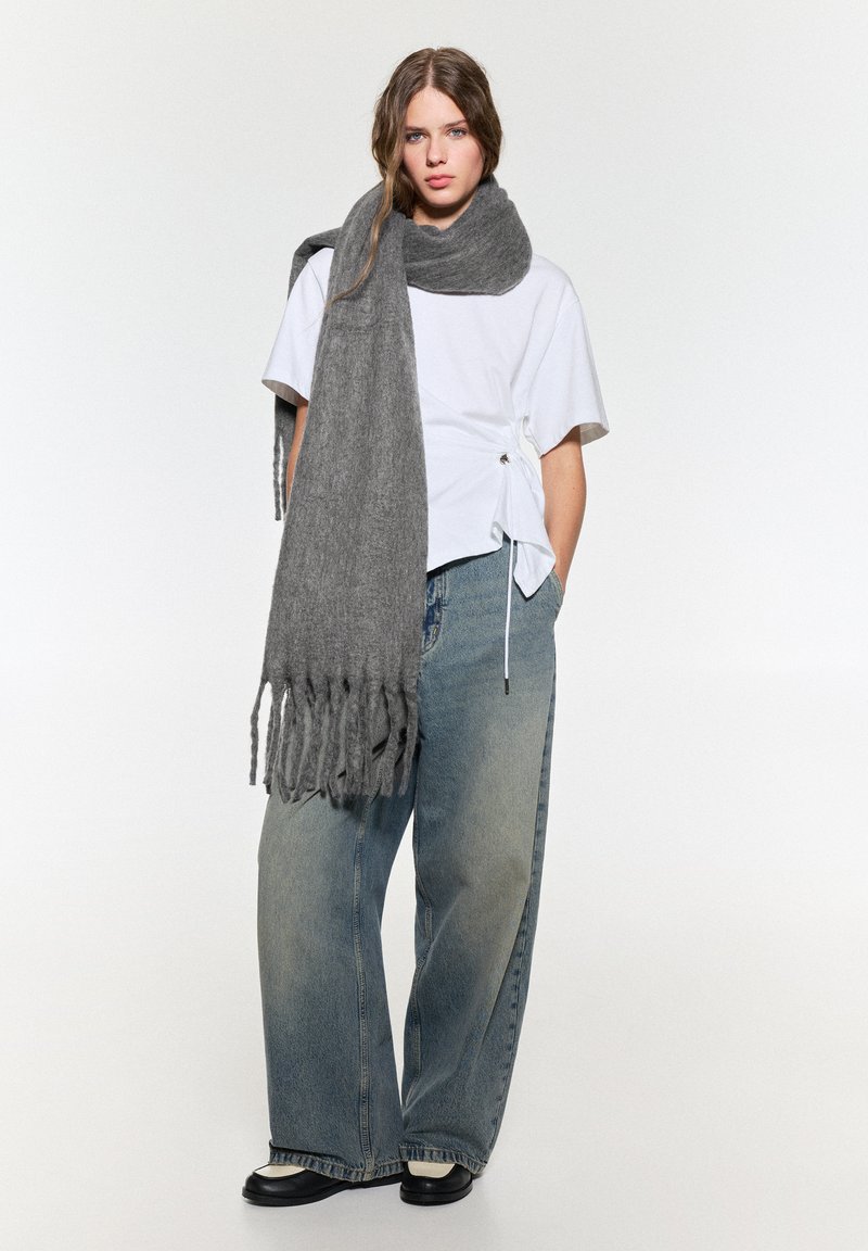 Grauer Strickschal mit Fransen, über einem weißen T-Shirt getragen. Das Model trägt lockere blaue Jeans und schwarze Schuhe und posiert vor einem neutralen Hintergrund.