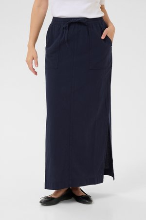 Jupe maxi bleu marine en tissu léger avec fente sur le côté, taille élastique et deux poches avant. Associée à des ballerines noires.