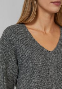 Vrouw in een grijs geribd trui met V-hals, die de nek, schouder en een deel van lichtbruin haar laat zien.