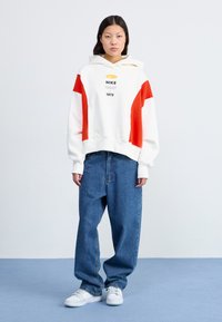 Hoodie blanc avec des accents rouges, présentant le logo Nike et le texte 1972. Associé à un jean bleu ample et des baskets blanches. Fond uni.