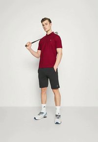 Camisola polo vermelha de mangas curtas, com logótipo no peito, combinada com calções pretos. O modelo segura um taco de golfe, vestindo sapatos e meias brancas de desporto.