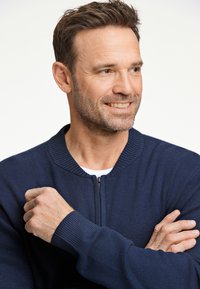 Marinblå zip-up tröja med ribbad krage och muddar, med ett texturerat stickmönster. Modellen har armarna i kors och ler.