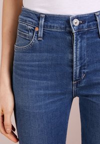 Jeans de mezclilla azul de cintura alta con un acabado suave. Presentan costuras visibles, un cierre de botón en la parte delantera y un diseño clásico de cinco bolsillos.