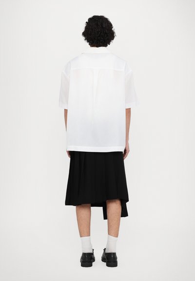 Weißes kurzärmeliges Hemd mit lockerer Passform, in einen schwarzen Faltenrock gesteckt. Schwarze Schuhe und knöchellange weiße Socken vervollständigen das Outfit.