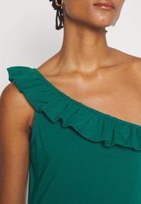 Femme portant un haut vert sans manches avec un col asymétrique à volants sur une épaule et une boucle d'oreille en or dans une oreille.