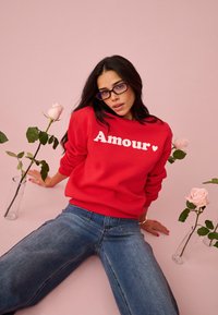 Femme portant un pull rouge "Amour" et des lunettes, assise sur un sol rose parmi des roses roses dans des vases en verre, avec un jean et de longs cheveux noirs.