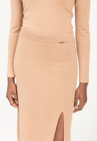 Gehaakte, beige jurk met lange mouwen en een textuur ribband in de taille. Heeft een zijsplit en een gouden accentdetail nabij de taille.