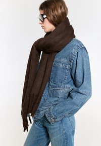 Écharpe en laine marron, texture douce, design long et épais avec des extrémités effilochées, enroulée autour d'une veste en denim de teinte bleue.
