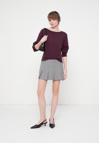 Burgundowy sweter z bufiastymi rękawami, szara plisowana mini spódnica oraz czarne buty na obcasie ze zapięciem na pięcie. Modelka trzyma małą czarną torebkę.