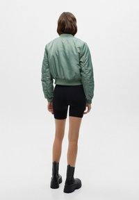 Groene bomberjack gemaakt van glad, glanzend materiaal met een geribbelde tailleband, gecombineerd met zwarte shorts en enkellaarsjes met dikke zolen.