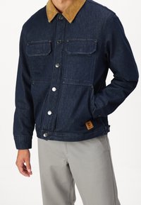 Tummansininen farkkutakki, jossa on ruskea samettikaulus, kahdella etutaskulla, nepparikiinnityksellä ja teksturoidulla pinnalla.