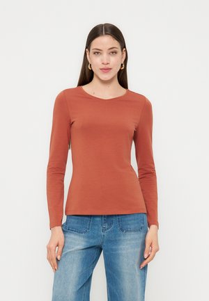 Mujer con camisa de manga larga color óxido y jeans azules, de pie frente a un fondo claro y liso, mirando hacia adelante.