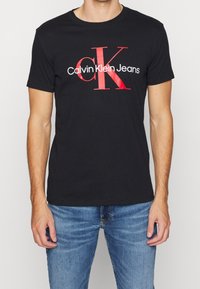 Svart bomulls t-shirt med en framträdande röd "CK" logo och "Calvin Klein Jeans" tryckt nedan i vitt. Korta ärmar och rund hals.