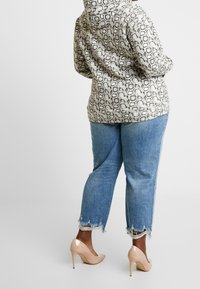 Osoba oblečená v mikině se vzorem hadí kůže, modré džíny s třepeným lemem a tělové vysoké podpatky, stojící na jednoduchém světlém pozadí.