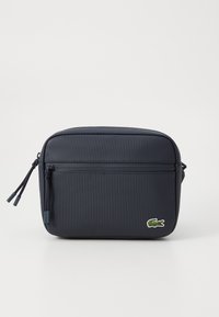 Geantă de umăr rectangulară de culoare bleumarin, confecționată din material sintetic texturat, având un buzunar frontal cu fermoar și un logo Lacoste verde.