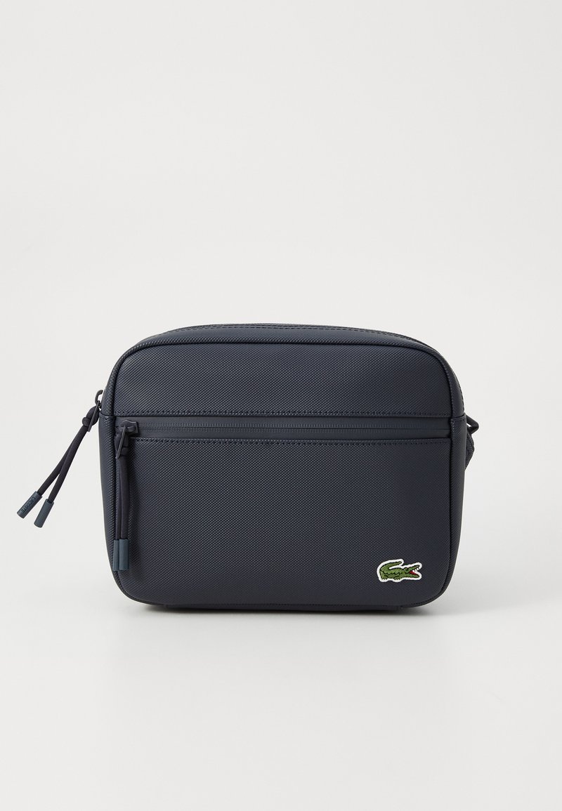 Geantă de umăr rectangulară de culoare bleumarin, confecționată din material sintetic texturat, având un buzunar frontal cu fermoar și un logo Lacoste verde.