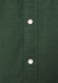 Tessuto di velluto a coste verde scuro con una trama a coste verticali e bottoni metallici a pressione a intervalli lungo la parte anteriore.