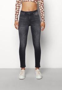 Mujer con jeans skinny negros desgastados de cintura alta, zapatillas blancas y un crop top marrón de manga larga con estampado de margaritas blancas, de pie frente a una pared blanca.