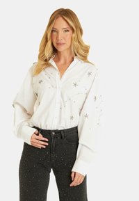 Camicia bianca a bottoni con decorazioni a forma di stella, abbinata a jeans neri con dettagli a pois argentati. Il tessuto appare morbido e testurizzato.