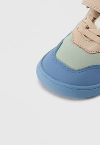 Αθλητικό παπούτσι με στρογγυλεμένη μύτη, διαθέτει μπλε λαστιχένια σόλα, μπεζ και πράσινο ανώτερο μέρος από δίχτυ, και υφή με ραμμένες λεπτομέρειες.