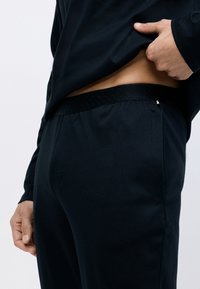 Pantalones deportivos negros con una cinturilla texturizada, bolsillos laterales y una tela lisa, mostrados de lado con una mano levantando ligeramente una camiseta.