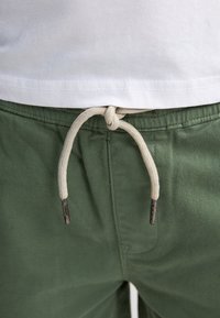 Shorts en coton vert avec une finition texturée, dotés d'un cordon beige noué et d'accents métalliques pour l'ajustement, associés à une chemise blanche.
