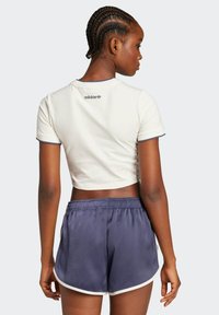 Bijela skraćena majica s tamnim obrubom i izvezenim logotipom, uparena s tamnoplavim šortsevima s elastičnom trakom i bijelim detaljima.