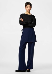 Haut noir à manches longues associé à un pantalon large bleu marine avec un devant tablier asymétrique et un détail noué à la taille.