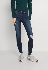 Skinny jeans in dark blue denim with distressed knee accent; paired with multicolored, sporty sneakers and a fitted green ribbed top.