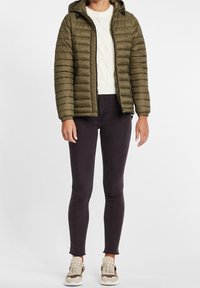 Veste matelassée vert olive avec design matelassé, fermeture éclair et capuche ajustable. Portée par-dessus un pull blanc en tricot et un jean skinny noir.