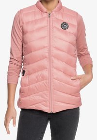 Roze, mouwloze gewatteerde bodywarmer met een chevron-patroon, hoge kraag, rits aan de voorkant en een logopatch. Gecombineerd met een roze top met lange mouwen eronder.