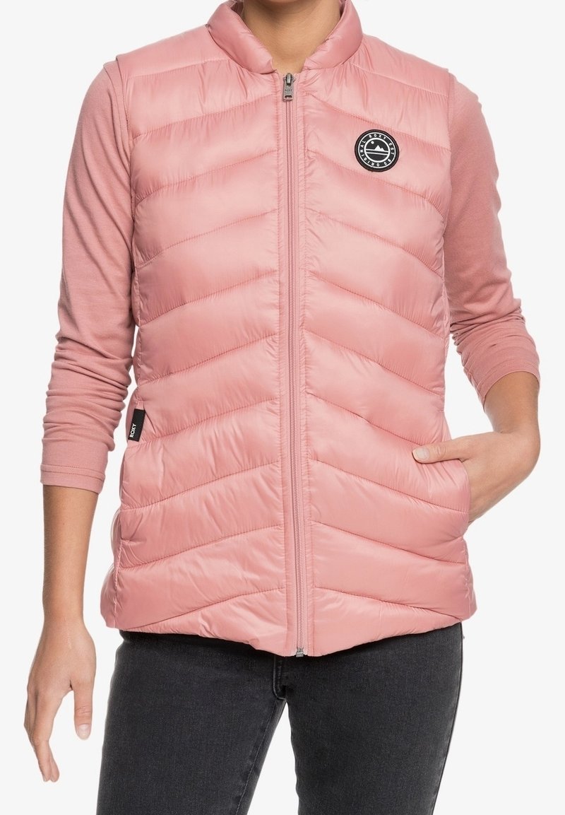 Roze, mouwloze gewatteerde bodywarmer met een chevron-patroon, hoge kraag, rits aan de voorkant en een logopatch. Gecombineerd met een roze top met lange mouwen eronder.