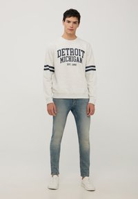 Jasnoszara bluza z napisem "Detroit Michigan", granatowe paski na rękawach, w połączeniu z zblakłymi niebieskimi dżinsami i białymi sneakersami.
