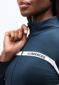 Κοντινό πλάνο ατόμου που κλείνει το φερμουάρ ενός σκούρου μπλε αθλητικού μπουφάν J.LINDEBERG με λευκή οριζόντια ρίγα στο ύψος του στήθους.