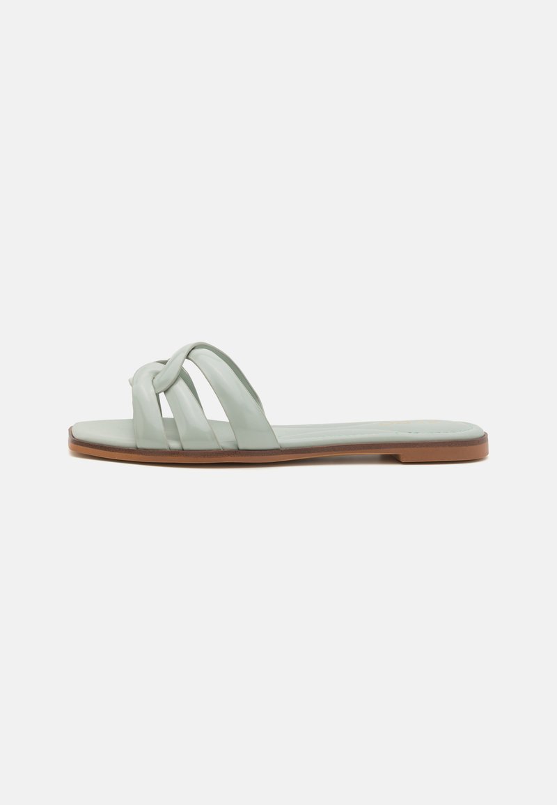 Sandales plates vert clair avec trois lanières croisées, une texture lisse et une semelle en caoutchouc marron. Design minimaliste sans embellissements.