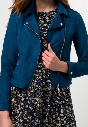 Femme portant une veste biker en daim bleu sarcelle sur une robe noire fleurie, tenant le revers de la veste avec la main droite.