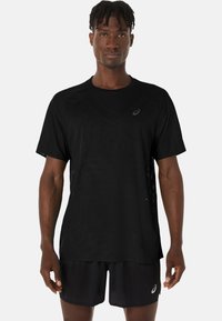 Schwarzere sportliche T-Shirt mit kurzen Ärmeln, das ein strukturiertes Stoffmuster und ein reflektierendes Logo auf der linken Brust aufweist. Getragen mit passenden Shorts.