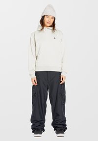 Lichtgrijze hoodie met een zak en logo, gecombineerd met zwarte baggy cargobroek met ritsen en zijzakken, gedragen met zwarte sneakers.