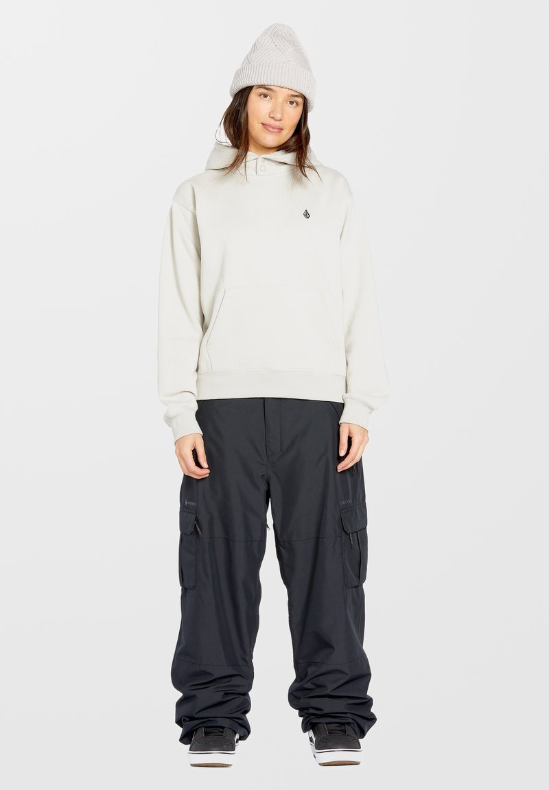 Lichtgrijze hoodie met een zak en logo, gecombineerd met zwarte baggy cargobroek met ritsen en zijzakken, gedragen met zwarte sneakers.