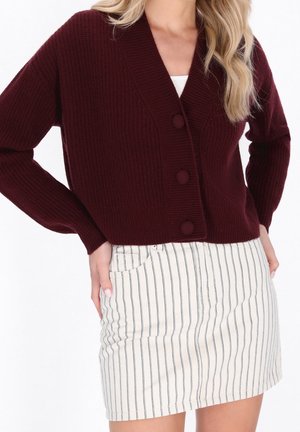 Femme portant un cardigan en tricot bordeaux avec de grands boutons et une mini-jupe blanche à rayures noires verticales.