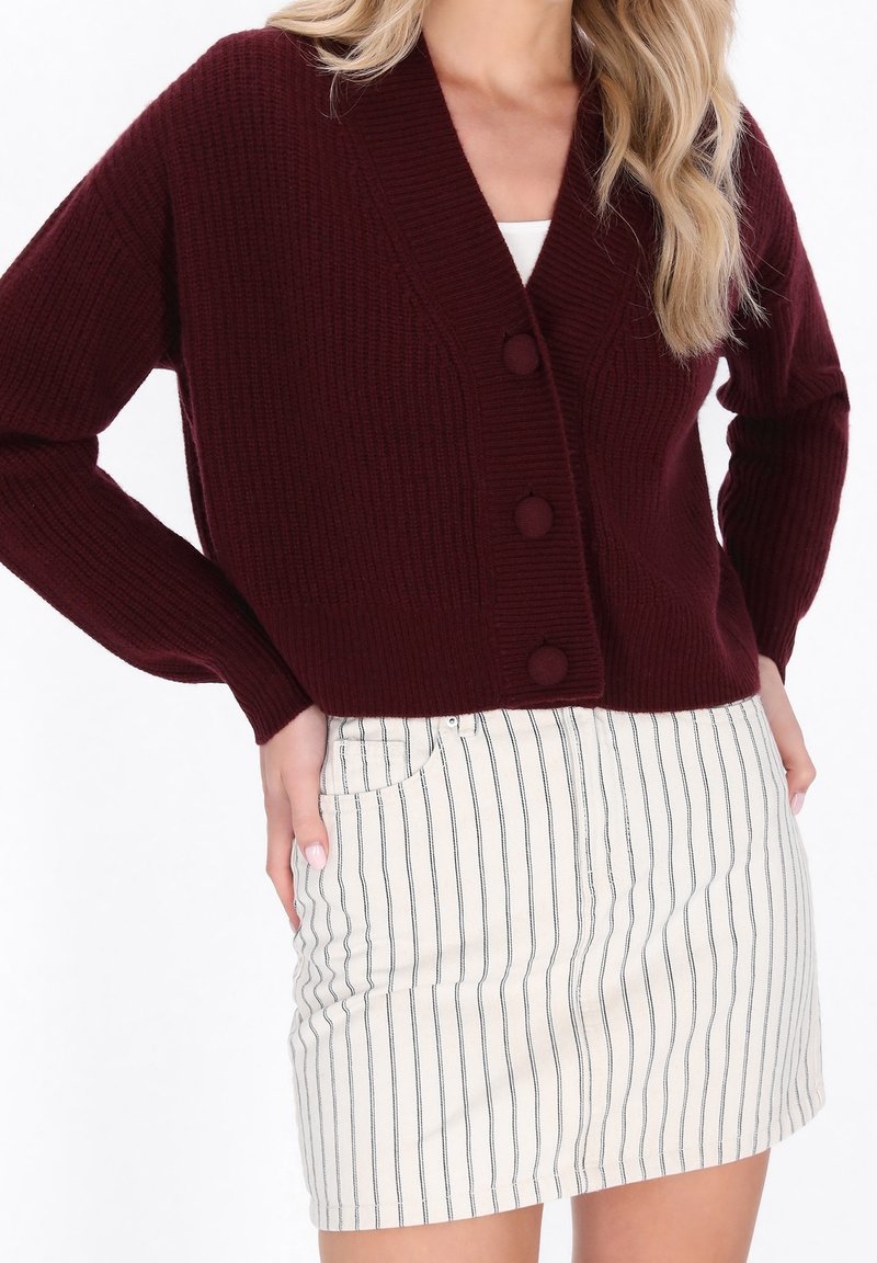 Femme portant un cardigan en tricot bordeaux avec de grands boutons et une mini-jupe blanche à rayures noires verticales.