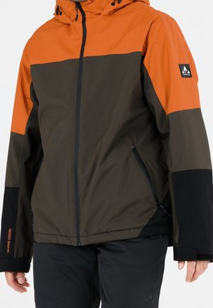 Person trägt eine Kapuzenjacke mit orangefarbenen, dunkelgrünen und schwarzen Farbfeldern, durchgehendem Reißverschluss vorne und seitlichen Reißverschlusstaschen, kombiniert mit schwarzen Hosen.