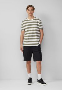 Olijf gestreepte t-shirt met een ronde hals, gecombineerd met zwarte cargo shorts. Model draagt zwarte sneakers en witte sokken. Neutrale achtergrond.
