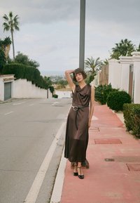 Femme en robe marron sans manches et chaussures noires posant sur un trottoir à côté d'une rue avec des palmiers et des haies en arrière-plan.