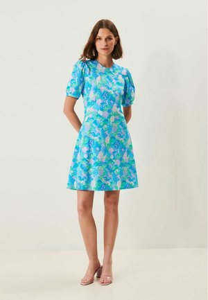 Kvinde iført en knælængde blå kjole med pufærmer og et flerfarvet blomsterprint, stående mod en ensfarvet lys baggrund.